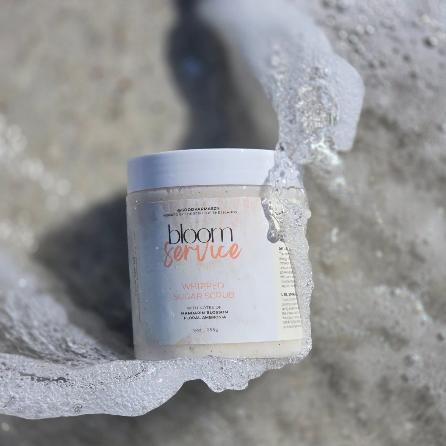 A container of whipped sugar scrub named Bloom Service  lays in the sand as water splashes around it.