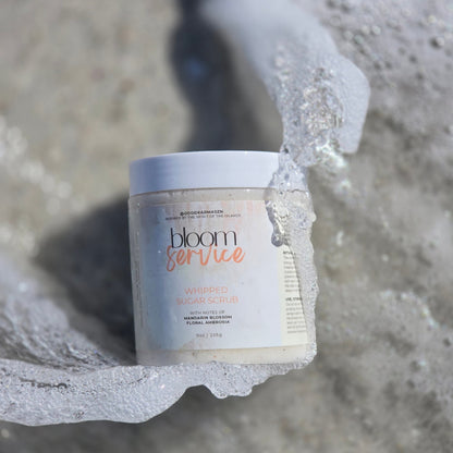A container of whipped sugar scrub named Bloom Service  lays in the sand as water splashes around it.