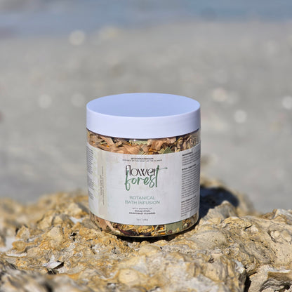 Jar of 'Flower Forest' botanical bath infusion on a rocky surface with a beach background