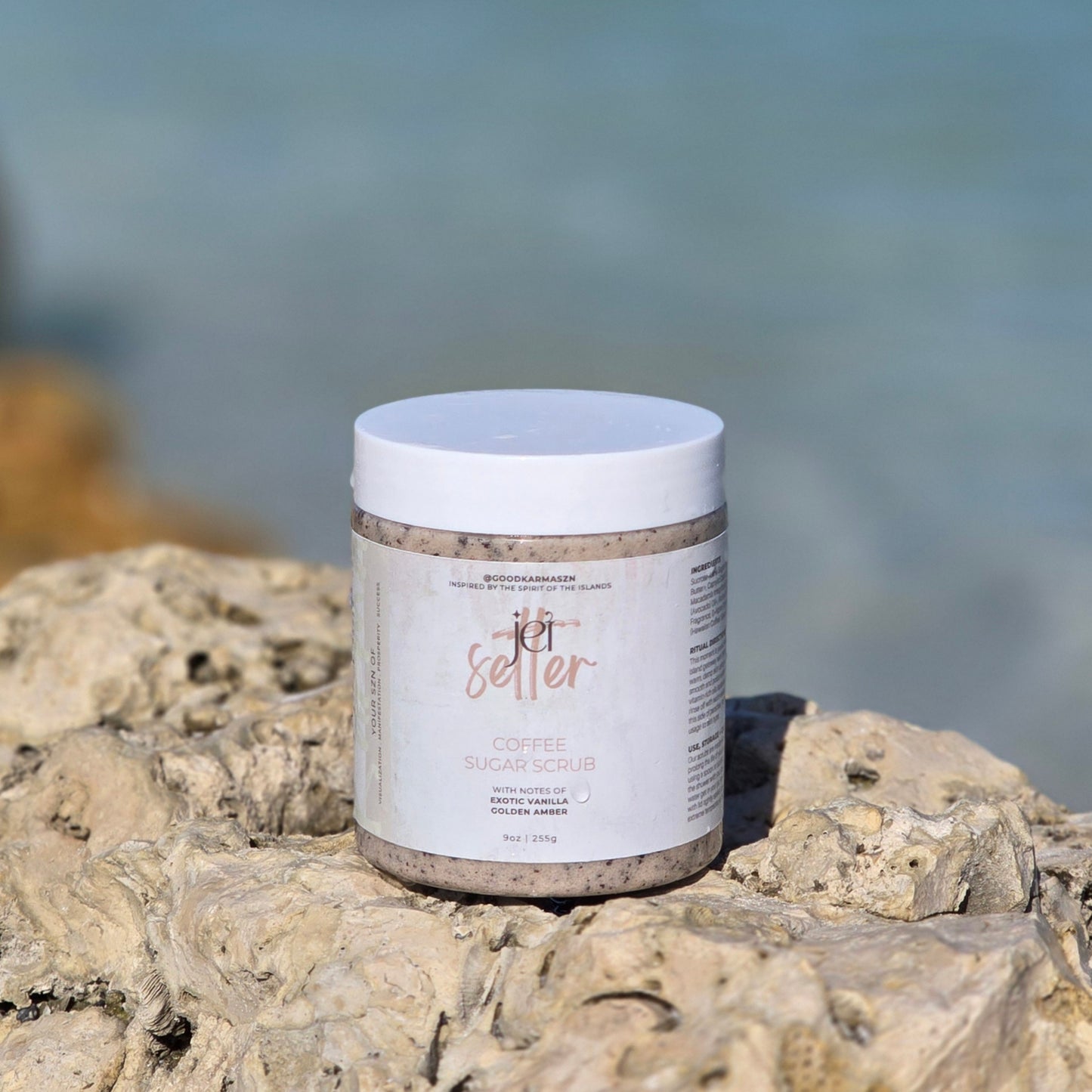 Container of a coffee and sugar scrub named Jet Setter on a rock with a blurred ocean background