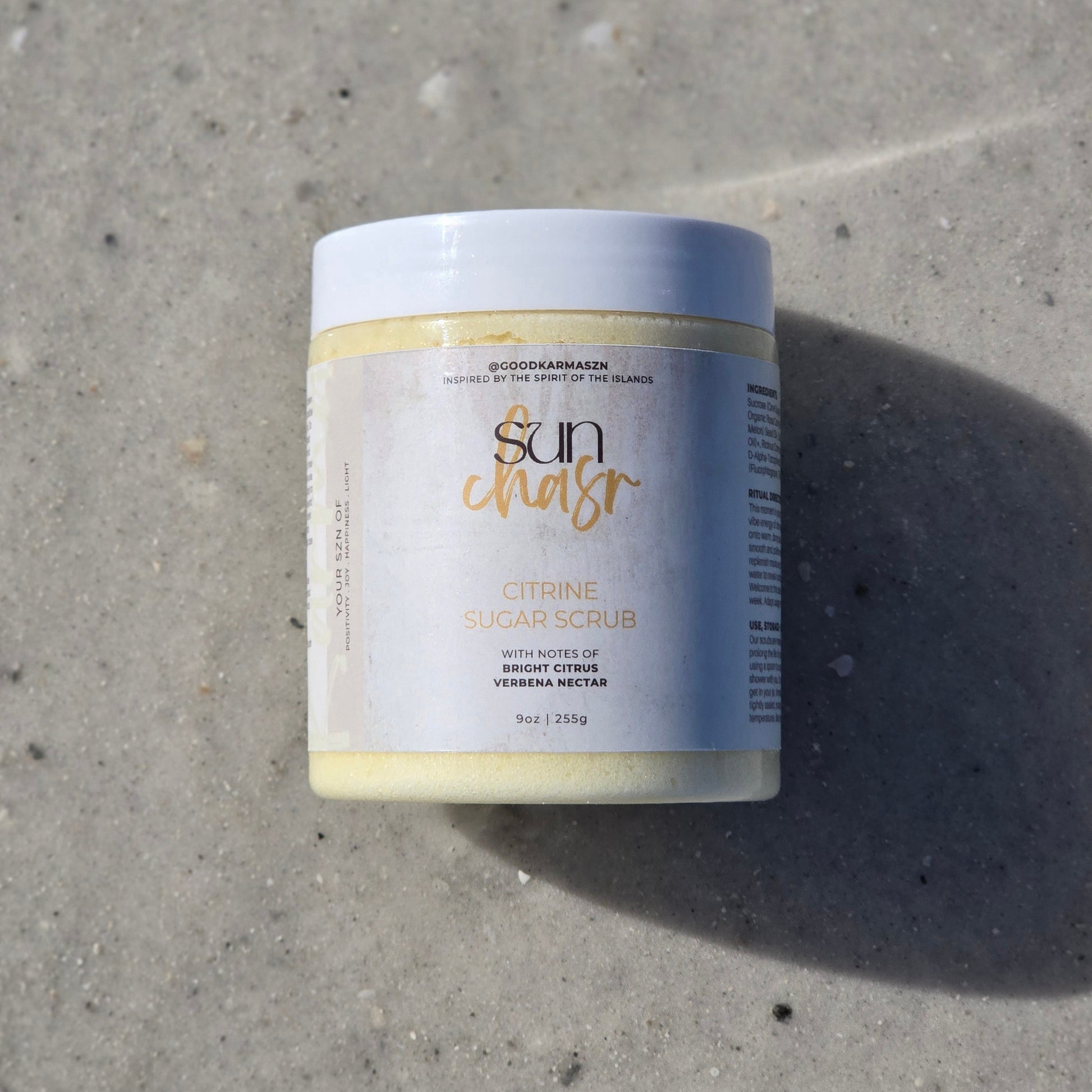 Container of whipped sugar scrub named Sun Chasr lays on the sand.